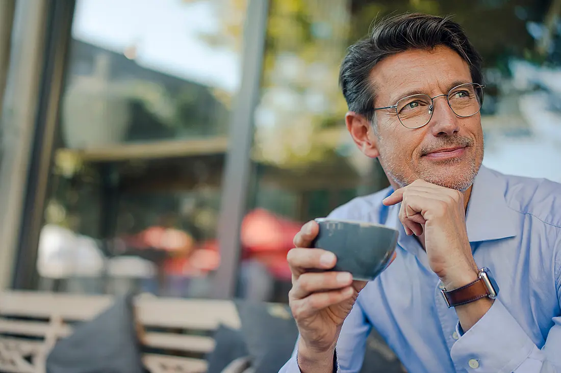 Ein Mann trinkt draußen eine Tasse Kaffee und schaut seitlich in die Kamera.