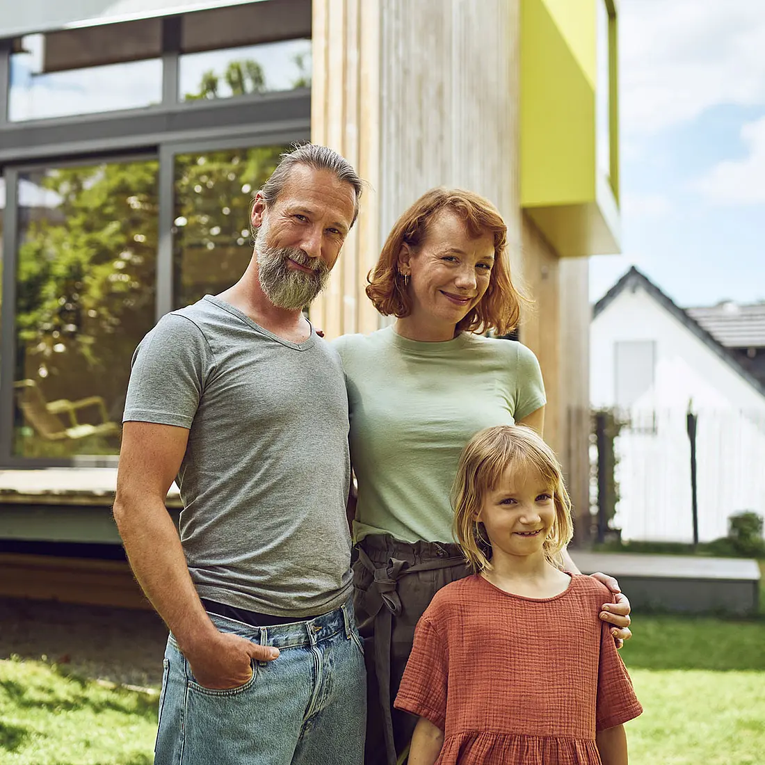 Junge Familie mit Vater, Muter und Tochter steht vor einem modernen Haus und schaut in die Kamera.