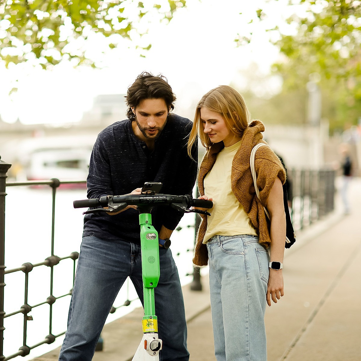 Junger Mann und junge Frau stehen am Ufer eines Flusses in einer Stadt und schauen gemeinsam auf ein Handy. Sie haben einen E-Roller dabei.