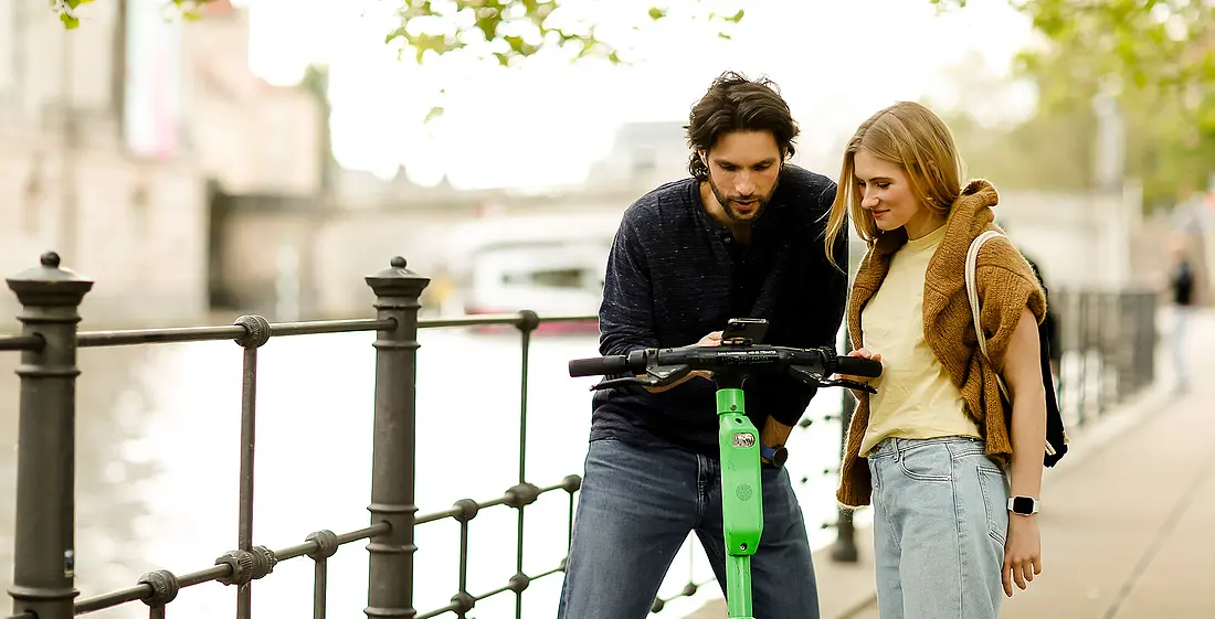 Junger Mann und junge Frau stehen am Ufer eines Flusses in einer Stadt und schauen gemeinsam auf ein Handy. Sie haben einen E-Roller dabei.