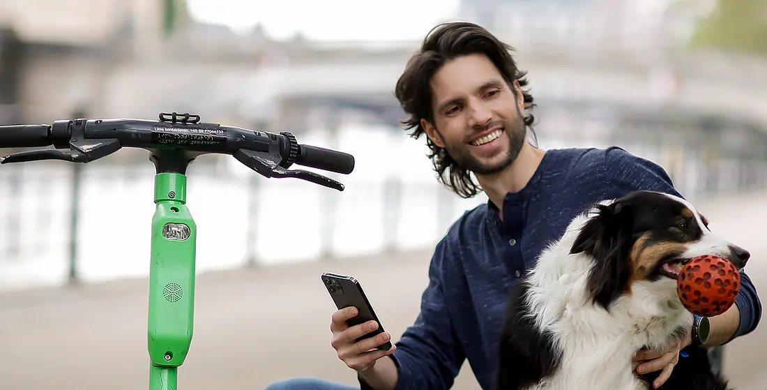 Ein junger Mann sitzt auf einer Mauer und tätschelt einen Hund. Neben ihm steht ein E-Roller.