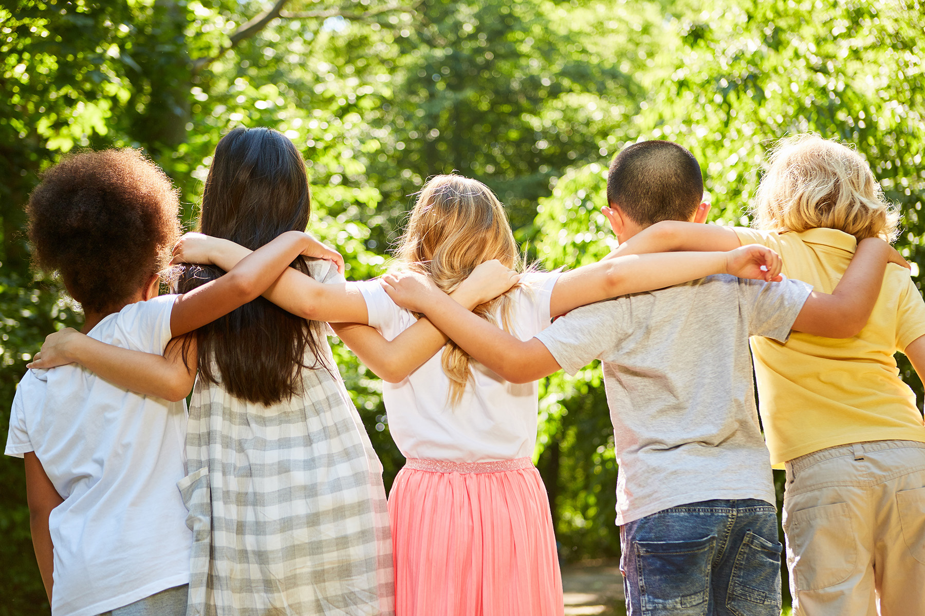 Gruppe von fünf Kindern steht mit dem Rücken zur Kamera. Sie haben sich gegenseitig die Arme um die Schultern gelegt.
